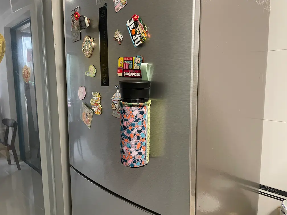 A colorful patterned magnetic neoprene bottle koozie stuck to a stainless steel refrigerator, highlighting vertical space-saving utility for offices and kitchens.