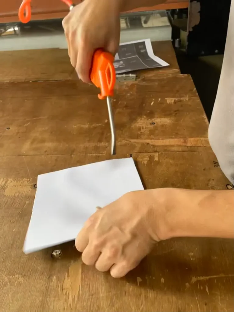 Hands using an orange air dust blower gun to clean fiber and dust from a white neoprene koozie blank panel on a wooden workbench. This critical step prevents fiber carbonization before sublimation.