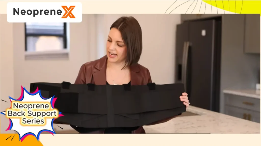 A female presenter holding up and demonstrating the unfolded black NeopreneX neoprene back support belt to show its double-layer tension bands and hook-and-loop fasteners in a bright indoor kitchen setting.