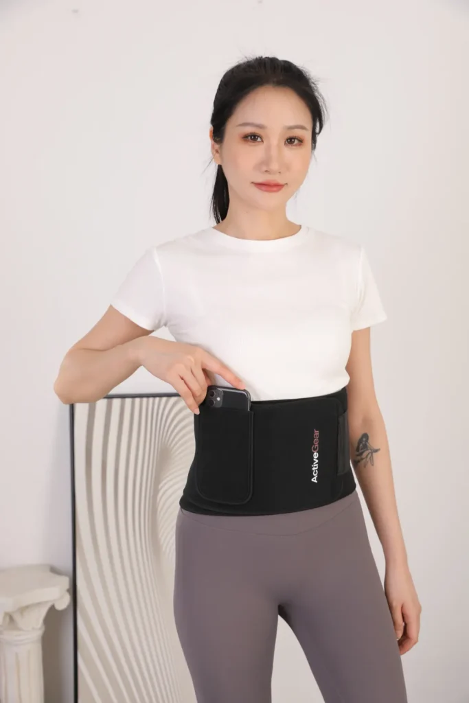 A fitness model inserting a smartphone into the built-in phone pocket of a black neoprene sweat belt, demonstrating functional pocket layout options for wholesale waist trimmers.